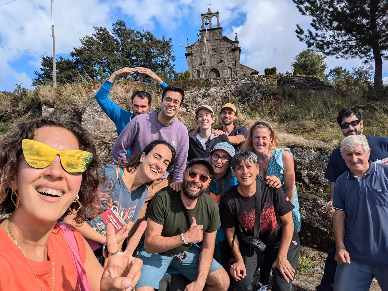 Selfie grupal dunha ducia de persoas nunha ladeira rochosa baixo unha pequena capela de pedra con espadana