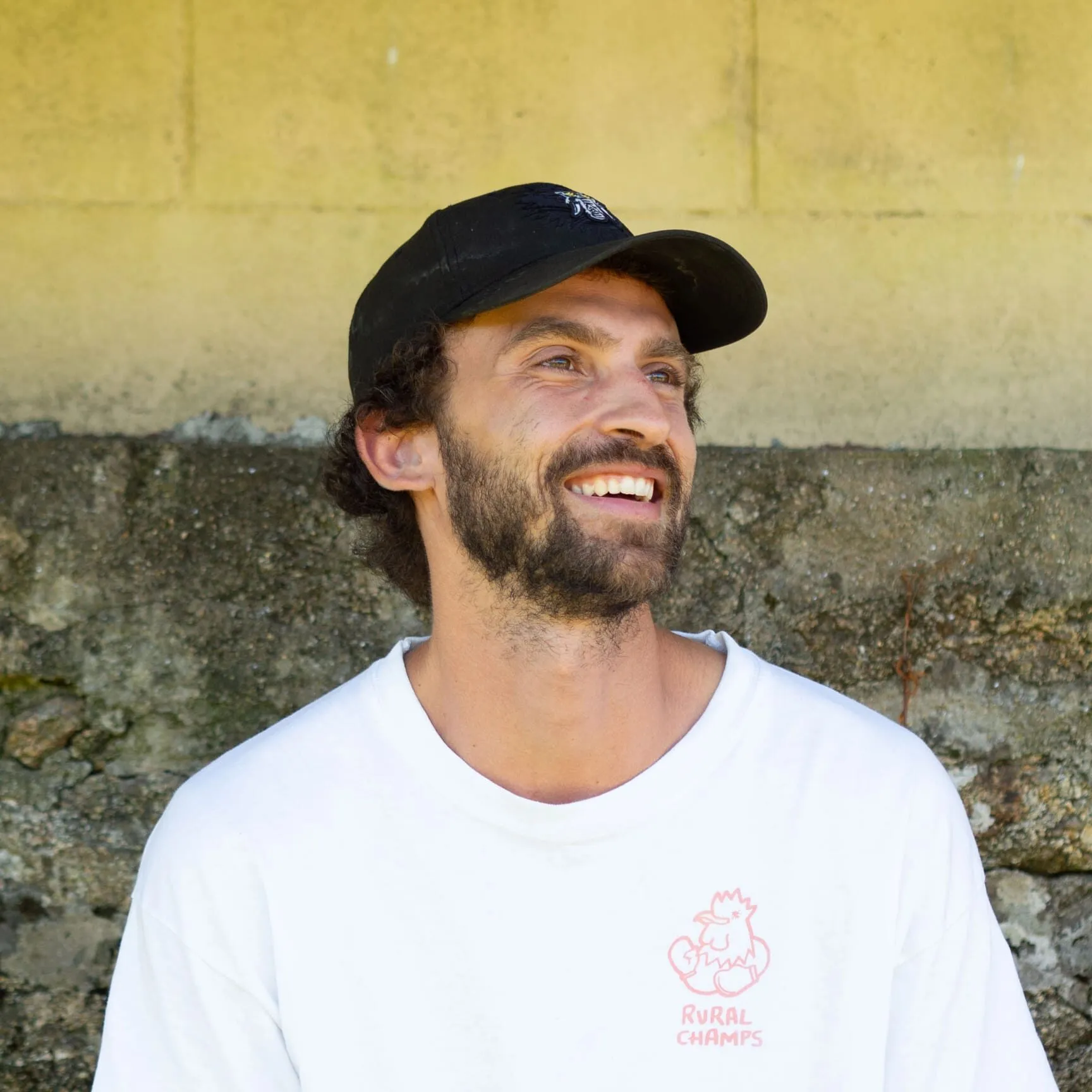 Víctor sonriendo con una gorra negra y una camiseta blanca de Rural Champs, apoyado en un muro de piedra amarillo