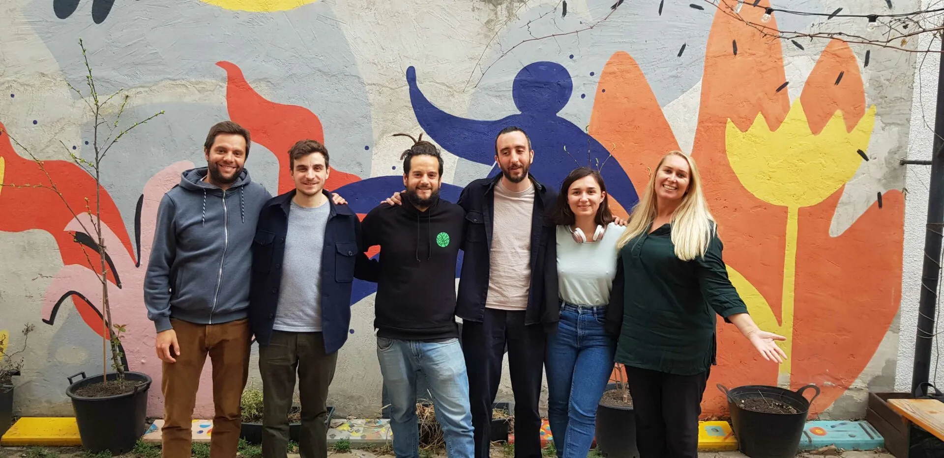 Seis persoas abrazadas nun patio, diante dun gran mural de cores cunha figura azul e flores