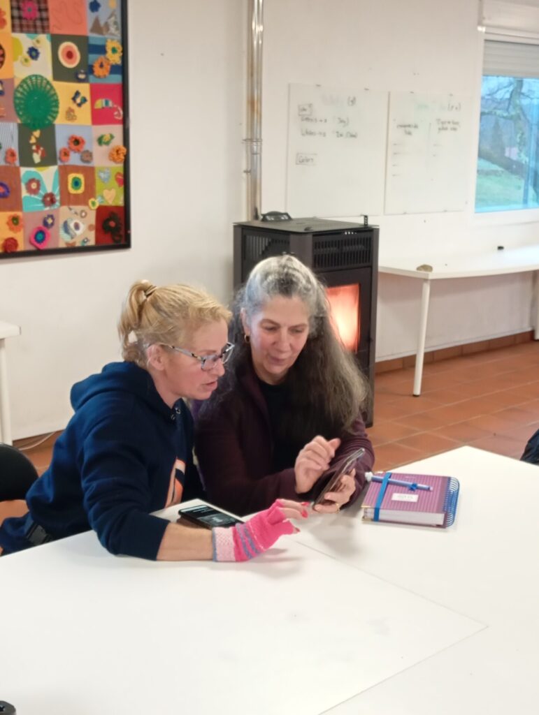 Two women sitting together at a table, one helping the other navigate a smartphone during a community session at Anceu Coliving