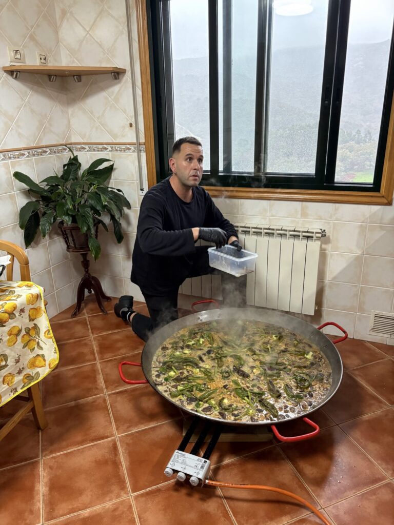A man cooking a large paella pan indoors at Anceu Coliving, with the green Galician hills visible through the window