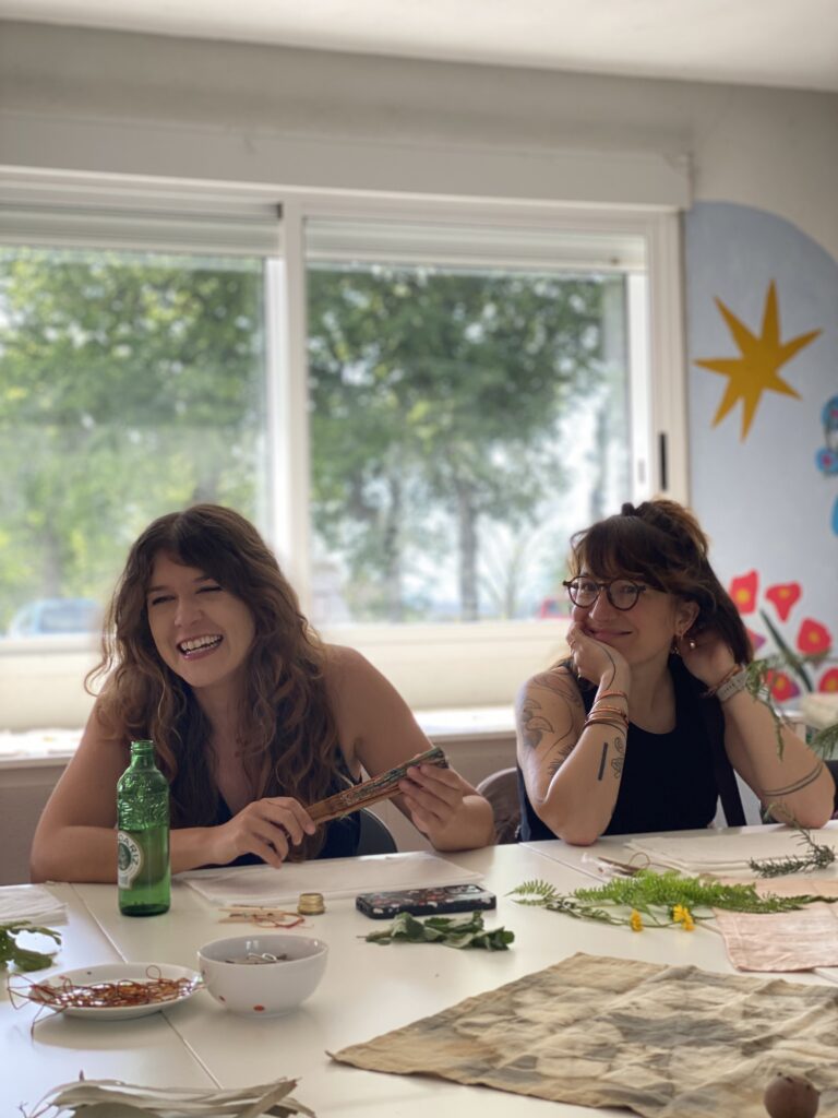 Two women laughing together during a creative workshop at Anceu Coliving, working on an artistic project with natural materials