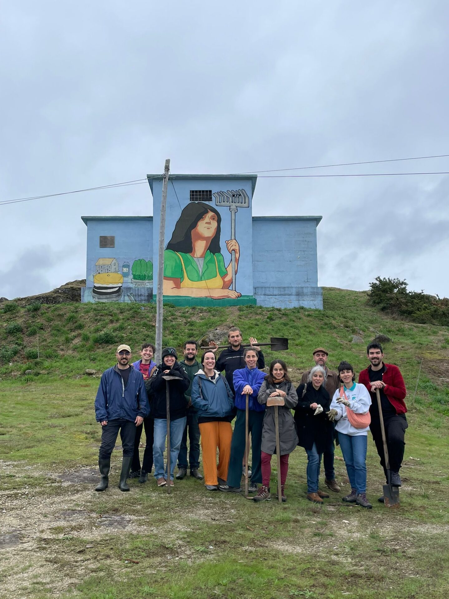 A group of people holding shovels and garden tools in front of a large painted mural on a hillside in rural Galicia, working on a community land project