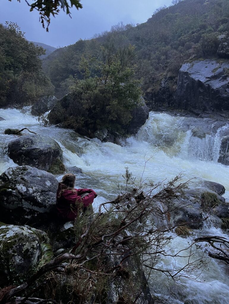 A person sitting on rocks beside a fast-flowing river surrounded by lush Galician forest on a rainy winter day